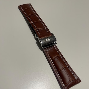 May include: A brown leather watch strap with a crocodile pattern and white stitching. The strap features a silver-colored clasp. The watch band is a replacement part for a watch.