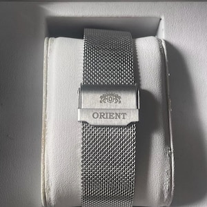 May include: A silver metal watch strap with a mesh design. The strap has a clasp with the word "ORIENT" engraved on it.