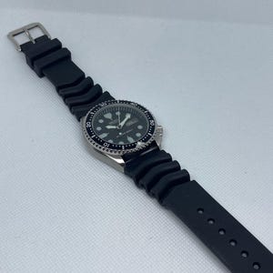May include: A black and silver wristwatch with a black rubber strap. The watch has a black face with white markings and a date window at the 3 o'clock position.