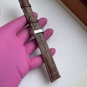 Peut inclure: Un bracelet de montre en cuir marron avec un motif crocodile et des coutures blanches. Le bracelet a une boucle argentée avec le nom de la marque. Le bracelet est tenu par une personne portant un gant rose.