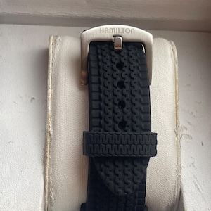 Peut inclure: Gros plan sur un bracelet de montre noir avec un motif texturé. Le bracelet est fait d'un matériau souple et est fixé à une boucle de couleur argent. Le mot "HAMILTON" est gravé sur la boucle.