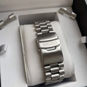 Könnte beinhalten: Ein silbernes Metallarmband mit der Gravur "SEIKO" auf der Schließe. Das Armband befindet sich in einer weißen Box mit schwarzem Rand.
