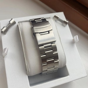 May include: A silver watch band with the word "SEIKO" engraved on the clasp. The band is in a white box with two small silver pieces.