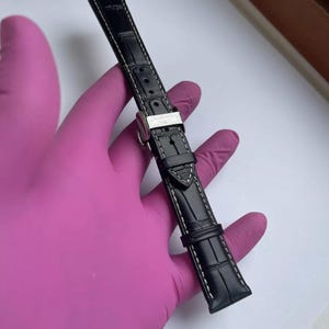 May include: Black watch strap with white stitching and a silver buckle. The strap has a textured, crocodile-like pattern. The buckle is engraved with the brand name.