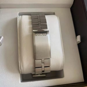 Puede incluir: Una correa de reloj plateada con la palabra "SEIKO" impresa en el cierre.