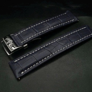 Puede incluir: Dos correas de reloj azul marino con costuras blancas y un patrón de cocodrilo. Una correa tiene una hebilla plateada. Las correas se muestran sobre una superficie oscura.