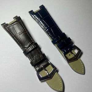 May include: Two watch straps with a glossy alligator-embossed texture. One strap is dark brown and the other is navy blue. Both feature a silver-toned metal buckle, two matching leather keepers, and a light beige underside with visible stitching.