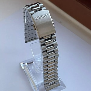 May include: A silver-toned Seiko watch band with a brushed metal finish. The band is displayed on a clear acrylic stand. The Seiko logo is visible on the clasp. The band is composed of multiple rectangular links.