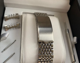 May include: A silver watch band with a brushed metal finish. The band is in a box with tools for adjusting the band.