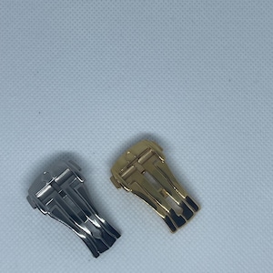 Puede incluir: Dos cierres de correa de reloj, uno plateado y otro dorado. Ambos cierres tienen un diseño similar con múltiples pliegues.