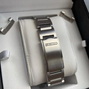 Puede incluir: Una correa de reloj de metal plateado con la palabra "SEIKO" grabada en el cierre.