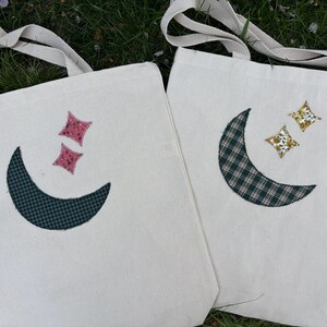 May include: Two white canvas tote bags with fabric appliques of moons and stars. One bag has a pink star and a green and white checkered moon. The other bag has a gold star and a green and white checkered moon.