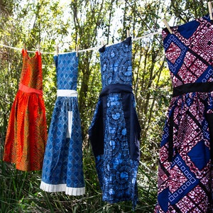 May include: Four colorful aprons hanging on a clothesline outdoors. The aprons are made of fabric with different patterns. One is orange, one is blue with white trim, one is blue with black trim, and one is red, white, and blue.
