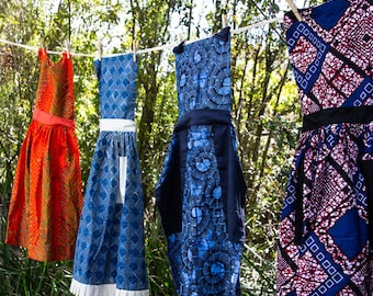 Könnte beinhalten: Vier bunte Schürzen hängen an einer Wäscheleine im Freien. Die Schürzen sind aus Stoff mit verschiedenen Mustern. Eine ist orange, eine ist blau mit weißem Besatz, eine ist blau mit schwarzem Besatz und eine ist rot, weiß und blau.