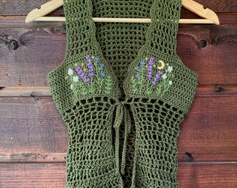 May include: Olive green crochet vest with embroidered lavender and moon details. The open-weave vest features a tie closure and is displayed on a wooden hanger against a wood-paneled background. The vest is a unique handmade garment.