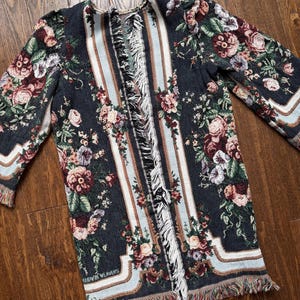 May include: A dark blue floral tapestry jacket with a fringed hem and vertical striped panels. The jacket features a repeating pattern of roses and other flowers in shades of pink, burgundy, and green. The sleeves are long.