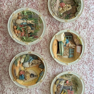 May include: Five circular decorative plates featuring three-dimensional relief scenes of rabbits in various settings. Each plate has a cream-colored border with small gold accents. The scenes depict rabbits in gardens, kitchens, and outdoor landscapes with detailed textures and colors.