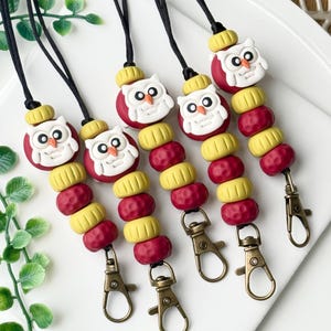 May include: Five lanyards featuring white owl-shaped beads, yellow cylindrical beads, and textured red spherical beads strung on black cords. Each lanyard ends with a bronze-toned metal lobster clasp for attaching items securely to the cord.