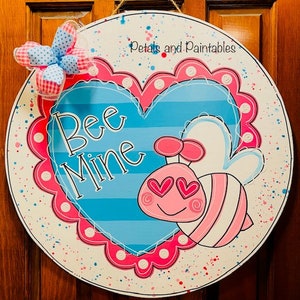 May include: Round wooden sign with a blue and pink heart design. The sign says "Bee Mine" and features a cartoon bee with heart-shaped eyes. A decorative bow is attached to the top left. The sign is painted with a white background and colorful accents.