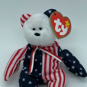 Puede incluir: Un oso de peluche patriótico con cabeza blanca y cuerpo con los colores de la bandera estadounidense. El cuerpo está dividido, con rayas rojas y blancas en un lado y estrellas blancas sobre tela azul en el otro. Lleva una etiqueta roja en forma de corazón.