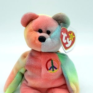 May include: A colorful plush bear with a peace sign embroidered on its chest. The bear features a tie-dye pattern in shades of pink, orange, yellow, green, and blue. It has a black nose and a tag attached to its ear.