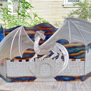 Może przedstawiać: Metalowa rzeźba smoka z rozpostartymi skrzydłami, osadzona na murze zamkowym. Rzeźba wykonana jest ze stali i ma srebrne wykończenie. Tło to niebieski i brązowy gradient.