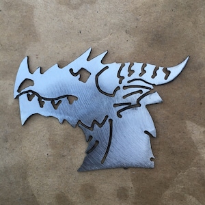 May include: A metal dragon head silhouette with intricate cut-out details. The dragon's profile features sharp teeth, a textured horn, and a detailed eye. The artwork is made of silver-colored metal.