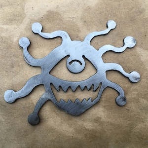 May include: A metal cutout of a monster with a single eye, sharp teeth, and multiple tentacles. The monster is silver in color and has a menacing expression.