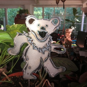 May include: A metal sculpture of a cartoon bear with a toothy grin, standing in a pot of soil and plants. The bear is painted in a silver colour and has a textured surface.