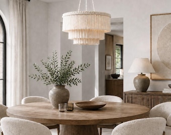 May include: A three-tiered cream-colored fringe chandelier hangs above a round wooden dining table. The table is surrounded by six chairs with light-colored textured upholstery and wooden frames, creating a cohesive dining room arrangement.