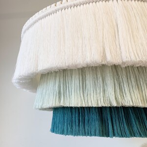 May include: A tiered, fringed lampshade in shades of white, green, and teal. The layers of fringe create a textured, decorative effect. The top layer is white, followed by a light green, and a deep teal at the bottom. The lamp is suspended from a white cord.