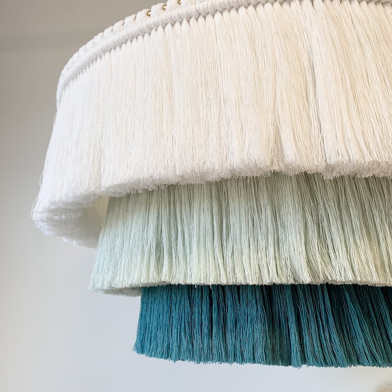 May include: A tiered, fringed lampshade in shades of white, green, and teal. The layers of fringe create a textured, decorative effect. The top layer is white, followed by a light green, and a deep teal at the bottom. The lamp is suspended from a white cord.