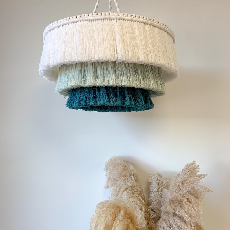 May include: A tiered, fringed chandelier in shades of white, seafoam green, and teal hangs from a white cord. The light fixture is suspended against a white wall, with dried pampas grass in the foreground.