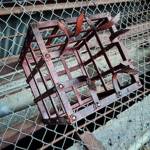 Puede incluir: Una jaula de metal oxidada con forma cuadrada sobre un fondo de malla de alambre. La jaula es de color rojo oscuro y está hecha de gruesos barrotes de metal.