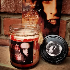 May include: A glass candle jar with a red wax design and a label that says "Interview with the Vampire". The jar is sitting on a brick surface next to a book with the same title. The book cover features a close-up of Tom Cruise's face.