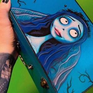 May include: A teal blue wooden box with gold accents and a hand-painted image of the Corpse Bride from Tim Burton's movie, "The Corpse Bride".