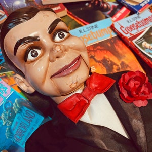 Pu&ograve; includere: Una bambola inquietante con un papillon rosso e una rosa rossa appuntata sulla giacca del suo vestito. La bambola &egrave; sdraiata su una pila di libri con il testo "Goosebumps" e "Horrorland" visibile.