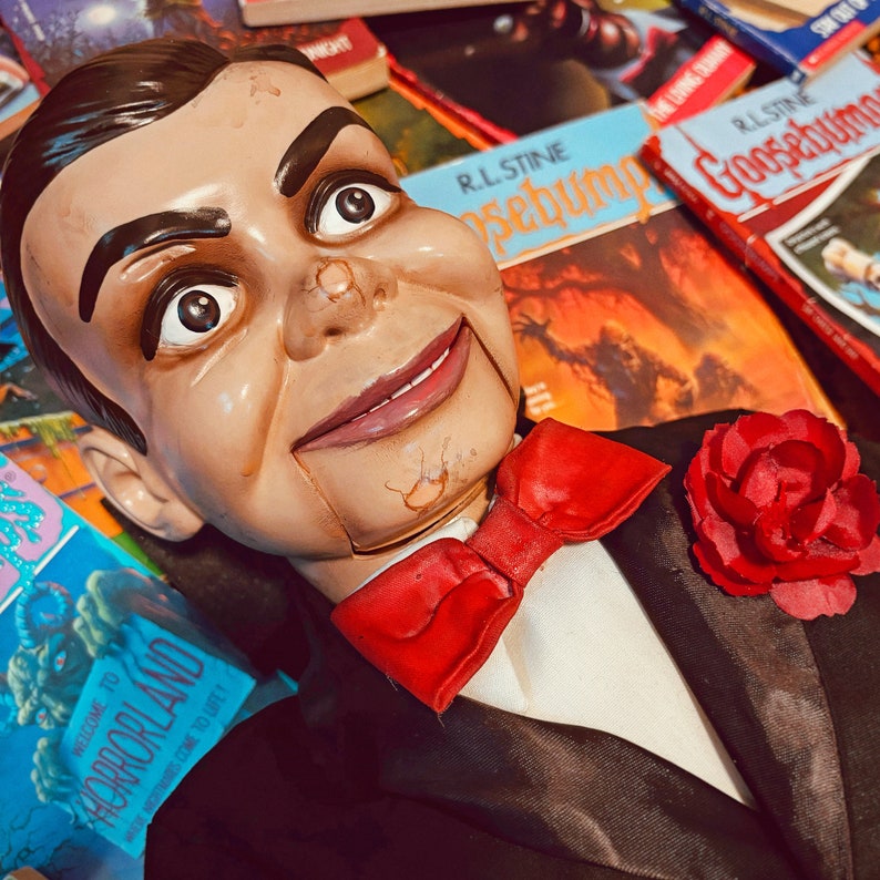Pu&ograve; includere: Una bambola inquietante con un papillon rosso e una rosa rossa appuntata sulla giacca del suo vestito. La bambola &egrave; sdraiata su una pila di libri con il testo "Goosebumps" e "Horrorland" visibile.