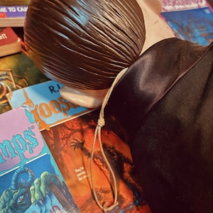 Pu&ograve; includere: Primo piano della testa di una bambola marrone con una lunga coda di cavallo marrone legata con uno spago beige. La bambola indossa una giacca nera. La bambola &egrave; sdraiata su una pila di libri tascabili con il testo "R.L. Stine Goosebumps" visibile su uno dei libri.
