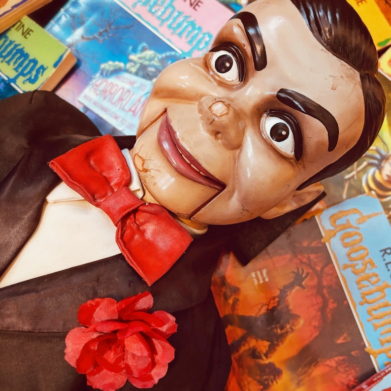 Pu&ograve; includere: Una bambola inquietante con un papillon rosso e una rosa rossa sul risvolto, che indossa un abito nero. La bambola &egrave; circondata da libri tascabili horror vintage con titoli come "Creepshow" e "The Horrorland".