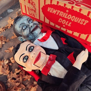 May include: Two vintage ventriloquist dolls with painted faces and black suits. The dolls are posed with their mouths open and their eyes looking up. The dolls are sitting on a red and white striped box with the words "Ventriloquist Doll" printed on it.
