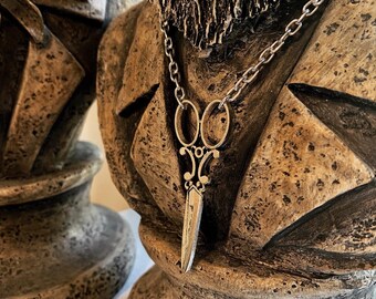 Op de afbeelding: Een zilverkleurige ketting met een gedetailleerde schaar hanger. De ketting wordt gedragen door een buste van een man met baard en een getextureerd, bruin oppervlak.
