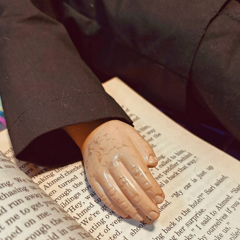 Pu&ograve; includere: Primo piano di una mano che tiene un libro aperto su una pagina con del testo. La mano &egrave; fatta di plastica ed &egrave; dipinta di un colore carne pallido. Il libro &egrave; bianco con testo nero. Il testo sulla pagina recita: "L'ho chiamato, venditore ambulante dietro, &egrave; tornato da quella parte."