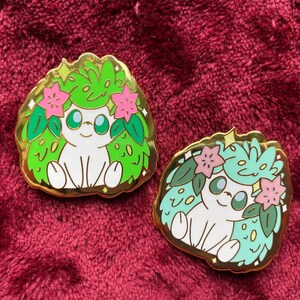 May include: Two enamel pins featuring a cartoon character with a white body, green and blue foliage, and pink flowers. The pins have a gold border and are set against a burgundy background. The character has large eyes and a smiling expression.