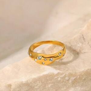 Puede incluir: Un anillo dorado con un dise&ntilde;o ondulado, engastado con m&uacute;ltiples piedras preciosas redondas y transparentes. El anillo se muestra sobre una superficie clara y texturizada, resaltando su acabado pulido y sus acentos brillantes. Una joya.
