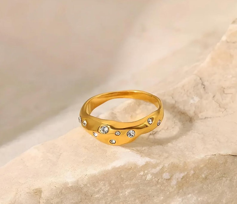 Puede incluir: Un anillo dorado con un dise&ntilde;o ondulado, engastado con m&uacute;ltiples piedras preciosas redondas y transparentes. El anillo se muestra sobre una superficie clara y texturizada, resaltando su acabado pulido y sus acentos brillantes. Una joya.