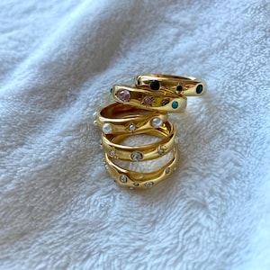 Op de afbeelding: Een stapel goudkleurige ringen met diverse versieringen. Sommige ringen hebben kleine, gekleurde edelstenen, terwijl andere parels en heldere kristallen hebben. De ringen zijn gestapeld op een wit, getextureerd oppervlak.