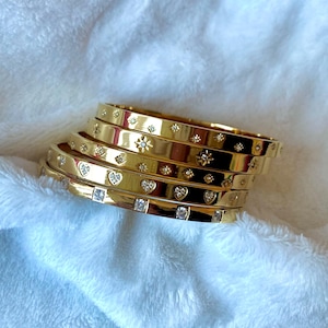 Puede incluir: Un conjunto de cinco pulseras doradas con diferentes diseños. Cada pulsera está adornada con pequeños diamantes de imitación transparentes en forma de estrellas, corazones y cuadrados.