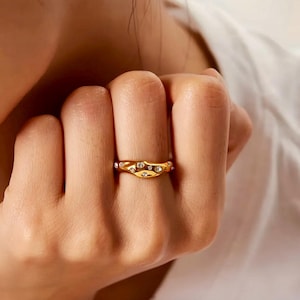 Puede incluir: Un anillo dorado con un dise&ntilde;o ondulado, adornado con m&uacute;ltiples piedras preciosas claras y brillantes. El anillo se lleva en un dedo, mostrando sus intrincados detalles y su estilo elegante. El dise&ntilde;o del anillo es llamativo y adecuado.