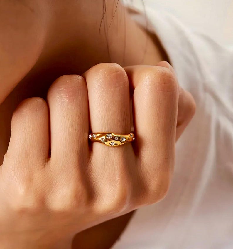 Puede incluir: Un anillo dorado con un dise&ntilde;o ondulado, adornado con m&uacute;ltiples piedras preciosas claras y brillantes. El anillo se lleva en un dedo, mostrando sus intrincados detalles y su estilo elegante. El dise&ntilde;o del anillo es llamativo y adecuado.